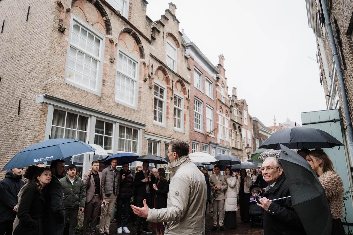 De gids vertelt een groep onder paraplu's in een straat met trapgevels