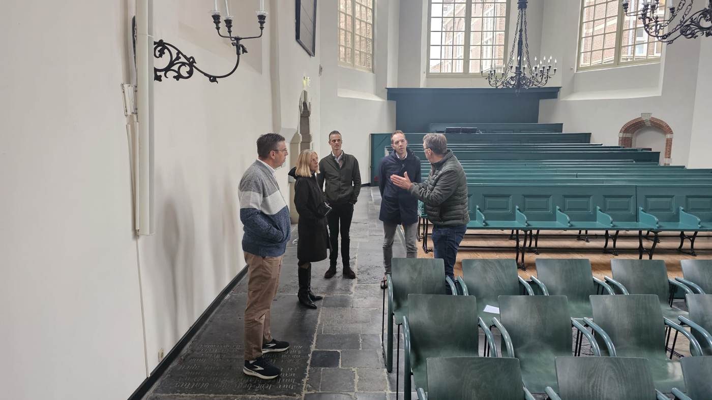Een groep in een witte kerkzaal met groene kerkbanken en kroonluchters
