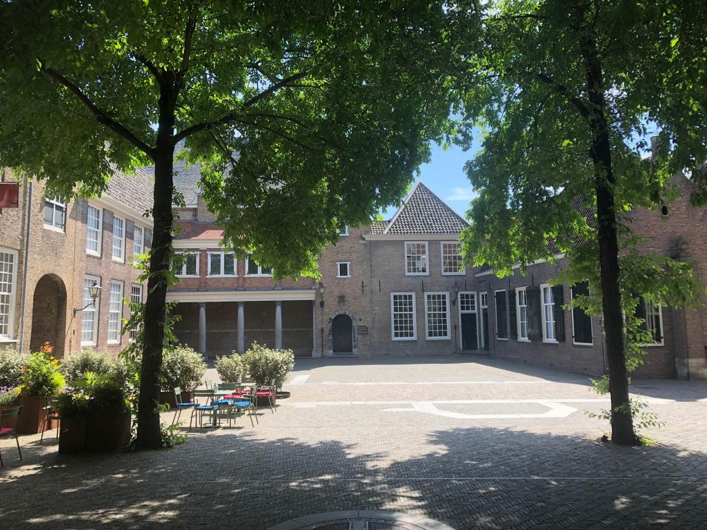 Een binnenplaats in de zon met bomen, een zuilengang en een terras met stoelen